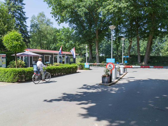 De ingang van Vakantiepark De Wielerbaan met de slagboom en het receptiegebouw, waar een fietser het park oprijdt.