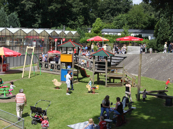 Overzicht van de centrale speeltuin op De Wielerbaan met glijbanen, schommels en andere speeltoestellen, naast een gezellig terras.