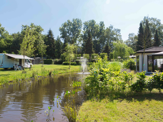 Een chalet en een caravan aan de rand van een vijver met fontein op het groene vakantiepark De Wielerbaan.