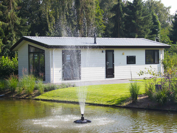 Een modern, wit chalet direct aan de waterkant op vakantiepark De Wielerbaan, met een fontein in de vijver voor de deur.