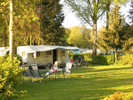 Ruime en zonnige kampeerplaats op het gras van vakantiepark De Wielerbaan, met een caravan, voortent en stoelen omgeven door bomen.