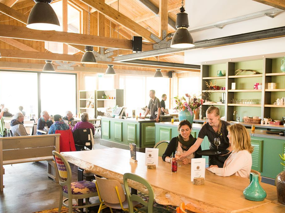 Gasten zitten aan een lange houten tafel in het sfeervolle, lichte restaurant van De Wije Werelt voor een drankje of maaltijd.
