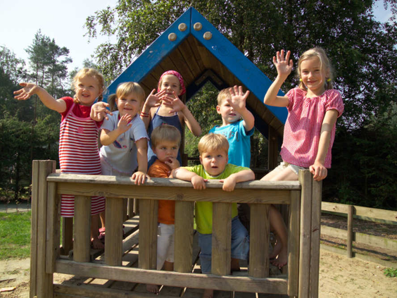 Een groep blije, zwaaiende kinderen in een houten speelhuisje in de buitenspeeltuin van vakantiepark De Wije Werelt op de Veluwe.