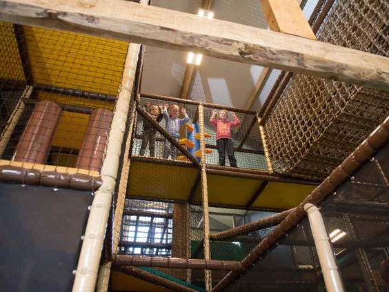 Kinderen zwaaien vanuit het hoge klim- en klauterparcours met netten in de overdekte binnenspeeltuin van De Wije Werelt.