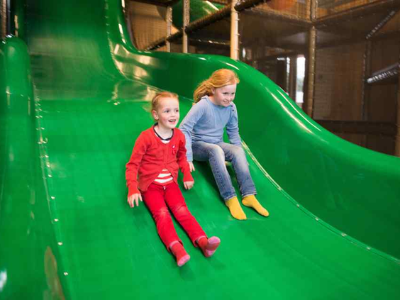 Twee meisjes glijden samen lachend van de grote groene glijbaan in de binnenspeeltuin van De Wije Werelt.