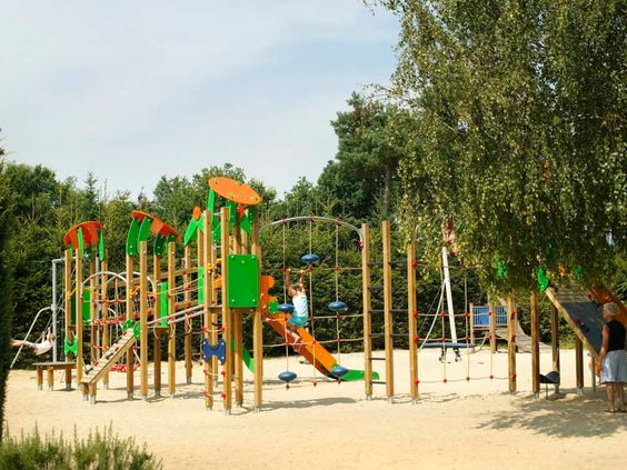 Grote buitenspeeltuin bij De Wije Werelt met houten klimtoestellen, een glijbaan en een grote zandbak, omringd door bomen.