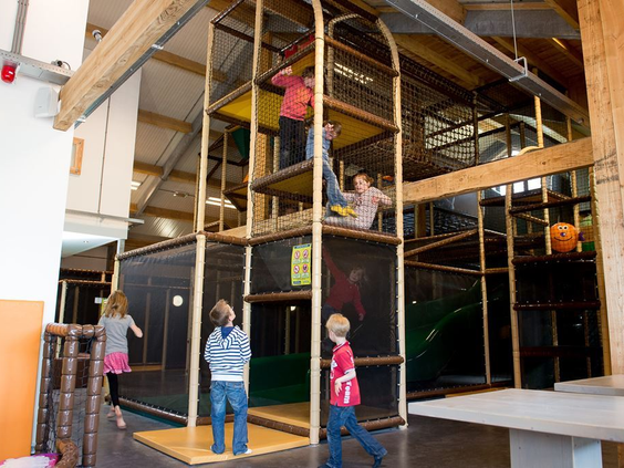 Kinderen klimmen en spelen in de grote overdekte binnenspeeltuin van De Wije Werelt, met meerdere verdiepingen, netten en glijbanen.