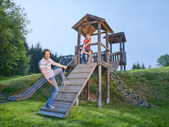 Een gezin met kinderen speelt samen in de natuurspeeltuin met een houten klimtoren en glijbaan op Dorint Vakantiepark Daun.
