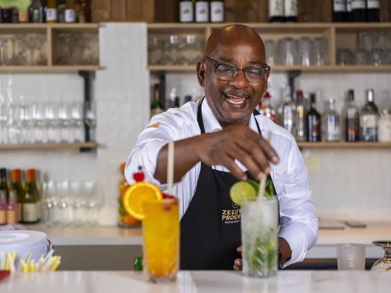 Een lachende bartender maakt kleurrijke cocktails en mocktails klaar achter de bar van het restaurant op Dormio Resort Nieuwvliet-Bad.