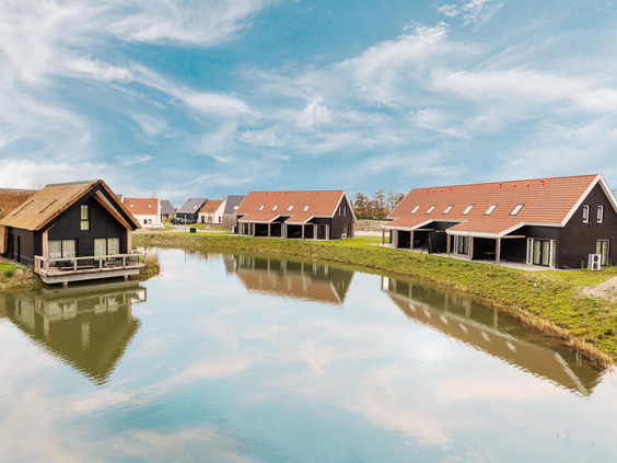 Moderne vakantiehuizen met rode daken aan het water op Dormio Resort Nieuwvliet-Bad, weerspiegeld in het water onder een blauwe lucht.