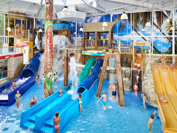 Overzicht van het kindergedeelte Playa in het Tikibad, met diverse brede, blauwe glijbanen, watersproeiers en spelende kinderen.