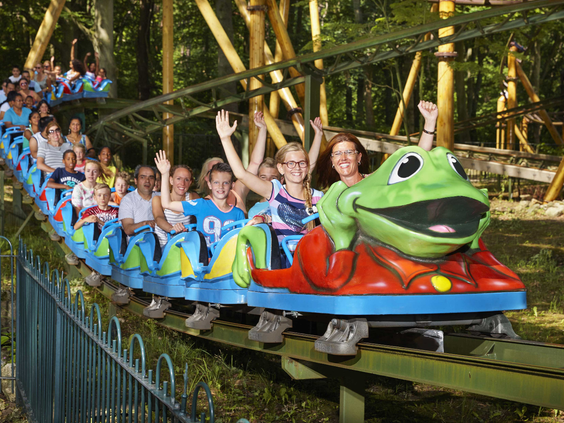 Een lange trein van de Kikkerachtbaan vol met lachende en zwaaiende families die genieten van de rit door het bos.