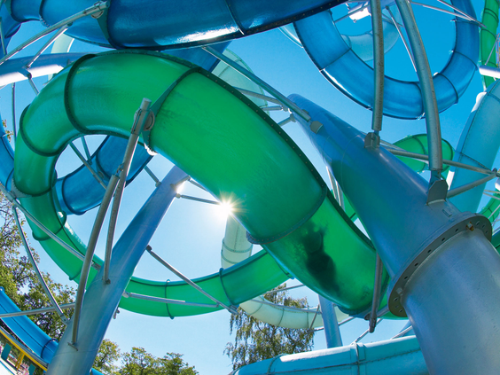 Een wirwar van groene en blauwe waterglijbaanbuizen van het Tikibad kriskras door elkaar, gefotografeerd van onderaf met de zon die erdoorheen schijnt.
