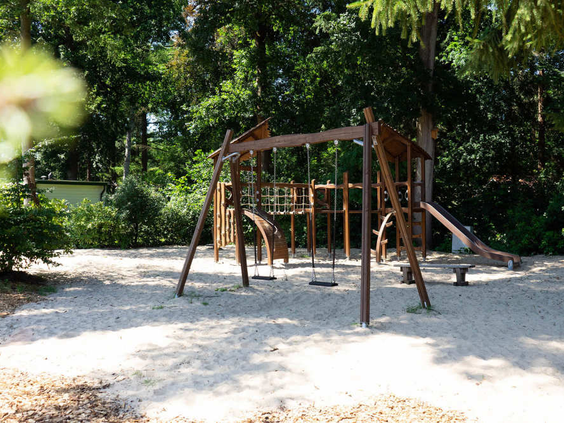 Een houten speeltuin met schommels en een klimtoestel op een ondergrond van zand, omringd door bomen.