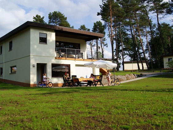 De buitenkant van een moderne vakantiewoning in Feriendorf Felsenhof, met een ruim grasveld en een terras waar gasten zitten.