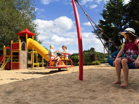 Kinderen spelen in de zonnige buitenspeeltuin van Feriendorf Felsenhof, met een glijbaan, schommels en een zandbak.