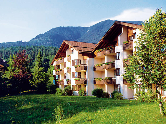 De buitenkant van het Dorint Sporthotel in Garmisch-Partenkirchen, omgeven door groene gazons en met de Beierse Alpen op de achtergrond.