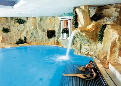Het luxe binnenzwembad van Dorint Sporthotel Garmisch-Partenkirchen met rotswanden en een waterval waar gasten onder relaxen.