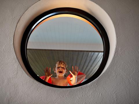 Een jongen met zwembandjes kijkt lachend door een patrijspoort in de speelruimte van Göbels Seehotel.