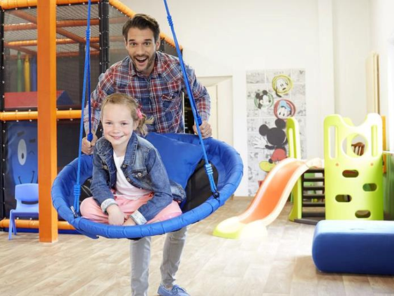 Een vader geeft zijn lachende dochter een duw op de grote schommel in de indoor speelruimte van Göbels Seehotel Diemelsee.