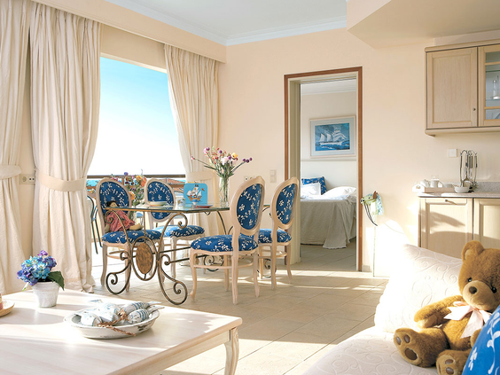 De ruime en lichte woonkamer van een familiesuite bij Grecotel Club Marine Palace, met een eethoek, een kitchenette en een doorkijk naar de aparte slaapkamer.