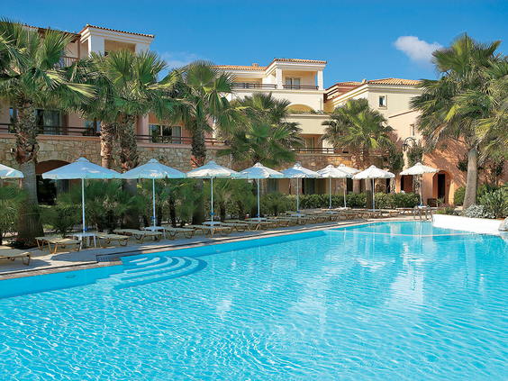 Een van de grote, blauwe zwembaden van Grecotel Club Marine Palace, omringd door ligbedden met witte parasols en de sfeervolle hotelgebouwen.