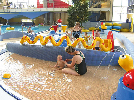 Een baby speelt met moeder in het ondiepe kinderbad van het Brockenbad, met een gele waterslang en andere speelelementen.