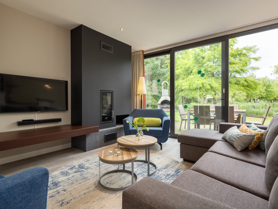 De stijlvol ingerichte woonkamer van een vernieuwde cottage in Het Heijderbos, met een moderne zithoek, sfeerhaard en grote ramen met uitzicht op het terras en de natuur.