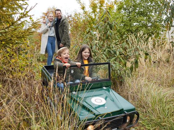 Een broer en zus rijden in een speelgoed-jeep tijdens de Kids Safari bij Center Parcs Het Heijderbos, terwijl hun ouders lachend toekijken.
