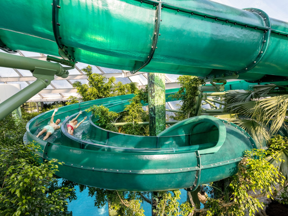 Een bezoeker glijdt met hoge snelheid door een lange, groene en deels doorzichtige waterglijbaan die tussen de tropische planten van Aqua Mundo slingert.