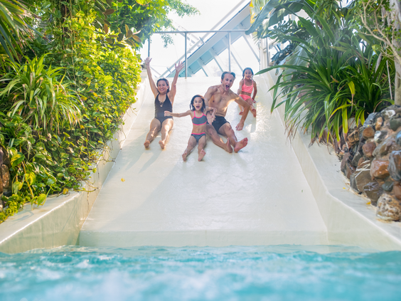 Een lachend gezin met drie kinderen glijdt hand in hand van de brede familieglijbaan in zwemparadijs Aqua Mundo van Center Parcs Het Heijderbos.