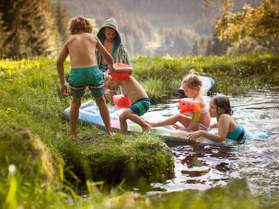 Vrolijke kinderen met zwemvleugels spelen in een ondiep beekje bij Hilserhof, waar ze dammetjes bouwen en de natuur ontdekken.