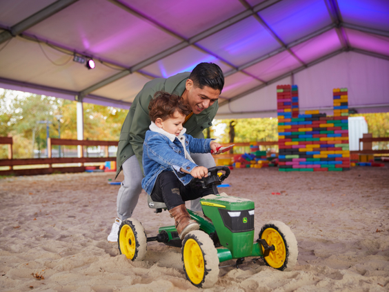 Een vader helpt zijn kind op een groene traptractor in de grote indoor zandbak van de Giga Konijnenhol speelhal op Hof van Saksen.