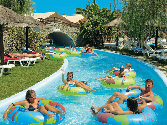 Gezinnen dobberen ontspannen in opblaasbanden op de lazy river van het Aqualand waterpark, omringd door groen en ligbedjes.