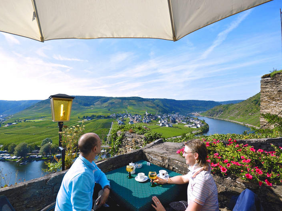 Twee volwassenen genieten van een drankje op het terras van Hotel Burgfrieden met een spectaculair panoramisch uitzicht over de kronkelende Moezelrivier en groene wijngaarden.