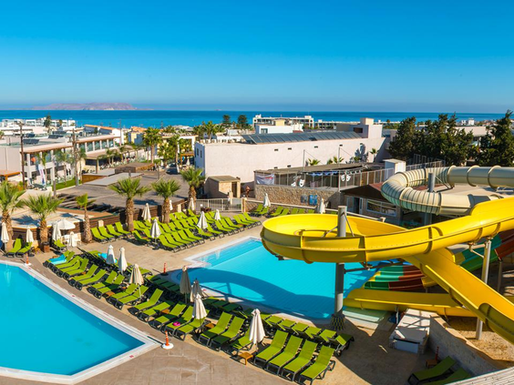 Zonnig overzicht van het waterpark met gele glijbanen en zwembaden, met de helderblauwe zee van Kreta op de achtergrond.