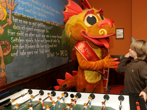 Een kind speelt tafelvoetbal met een vrolijke, gele drakenmascotte in de speelruimte van Hotel Ling Bao.