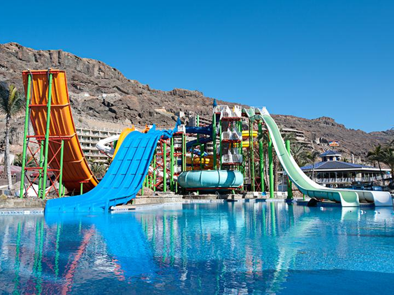 De hoge en kleurrijke waterglijbanen van het Aquapark Lago Taurito, met een brede blauwe en een steile oranje glijbaan die uitkomen in het zwembad.