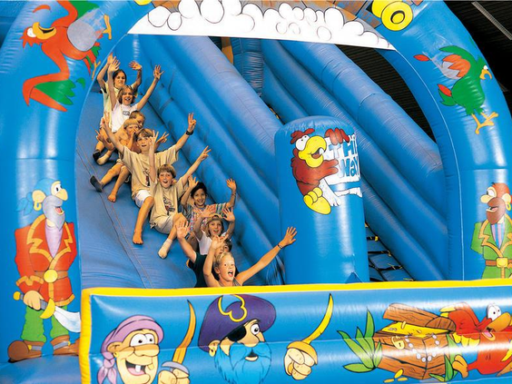 Een groep blije kinderen glijdt met de armen in de lucht van een groot blauw opblaasbaar piratenschip in het Kinderparadijs van Hotel Sauerland Stern.