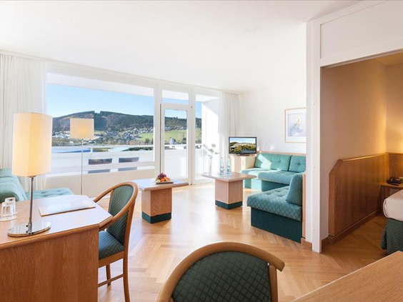 Ruim en licht appartement in Hotel Sauerland Stern met een aparte zithoek, bureau en een groot raam met uitzicht op de heuvels van Sauerland.
