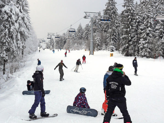 Kinderen en volwassenen skiën en snowboarden op een brede, besneeuwde piste in Winterberg, met een stoeltjeslift op de achtergrond.