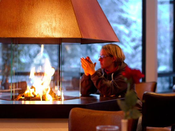 Een gast warmt zich op bij de gezellige, centrale open haard in de lounge van Hotel Winterberg Resort, met uitzicht op de sneeuw.