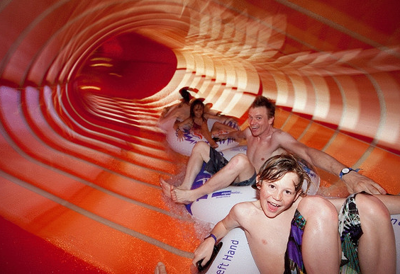 Een blij gezin glijdt samen in een grote opblaasband door een snelle, oranje waterglijbaan in het subtropische zwembad.