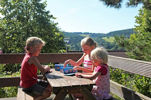 Drie kinderen doen een spelletje aan een picknicktafel op het balkon van hun vakantiehuis, met uitzicht over de heuvels van de Eifel.