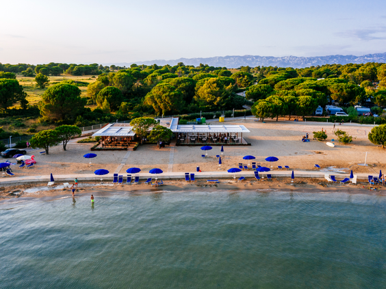 Luchtfoto van het kindvriendelijke zandstrand van Zaton Holiday Resort, met blauwe parasols en een beachbar aan de rustige Adriatische Zee.