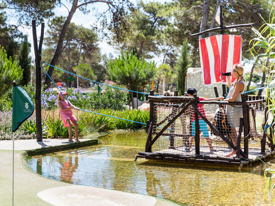 Kinderen steken met een houten trekvlot met piratenvlag het water over op de avontuurlijke golfbaan van Zaton Holiday Resort.