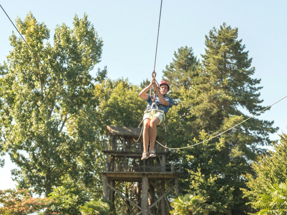 Kind zoeft van een hoge houten toren aan de zipline in het avonturenpark van de kindvriendelijke camping Domaine des Ormes.