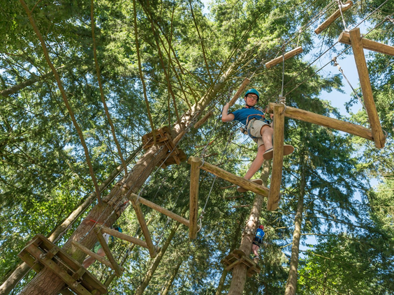 Kind met helm balanceert op een touwbrug hoog tussen de bomen in het klimbos van camping Domaine des Ormes.