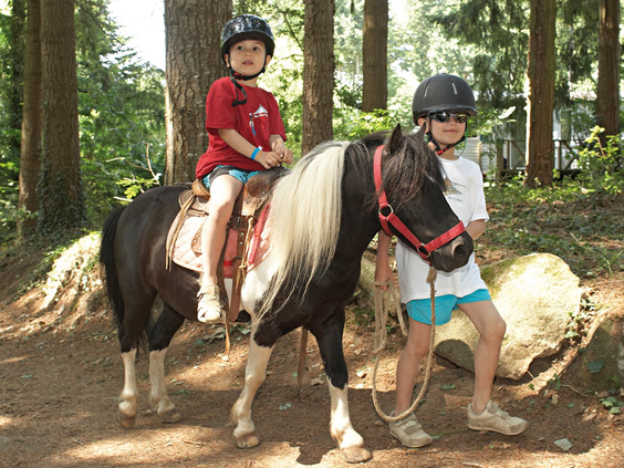 Kind met helm zit op een pony die wordt geleid door een ander kind door het bos op de kindvriendelijke camping Domaine des Ormes.