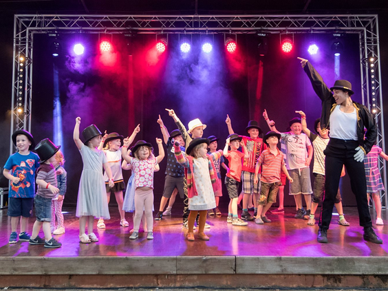 Groep kinderen staat op het podium te dansen en zwaaien met hoge hoeden tijdens een show op de kindvriendelijke camping Domaine des Ormes.